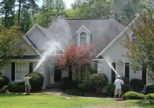 Two workers softwashing the front of a house, spray fanning across the pale siding