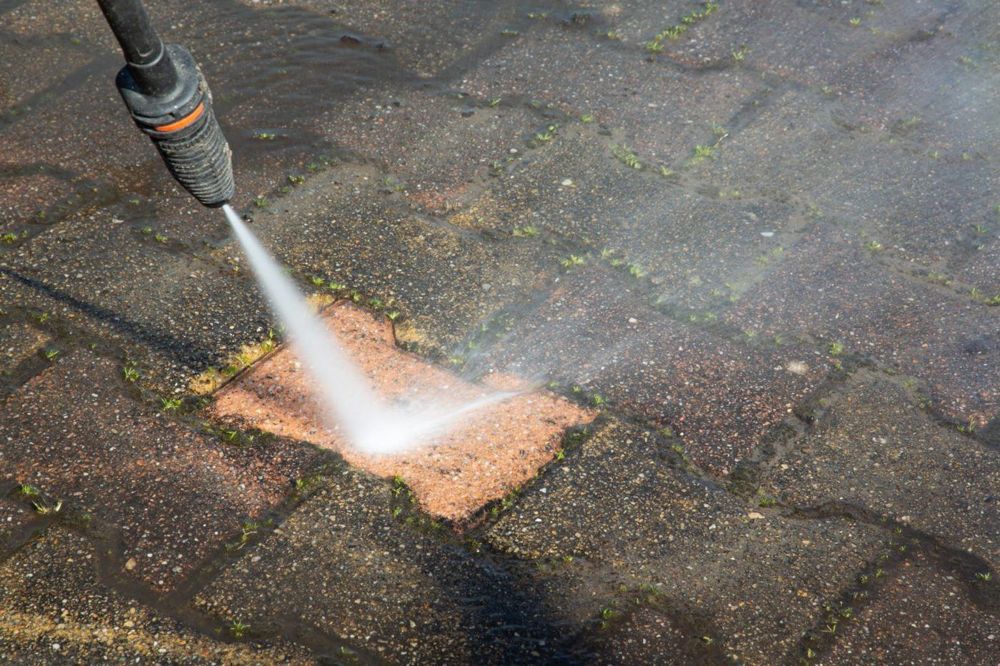 A pressure washer nozzle cleaning a paver, leaving one brick bright orange against the surrounding dirty, grey pavers
